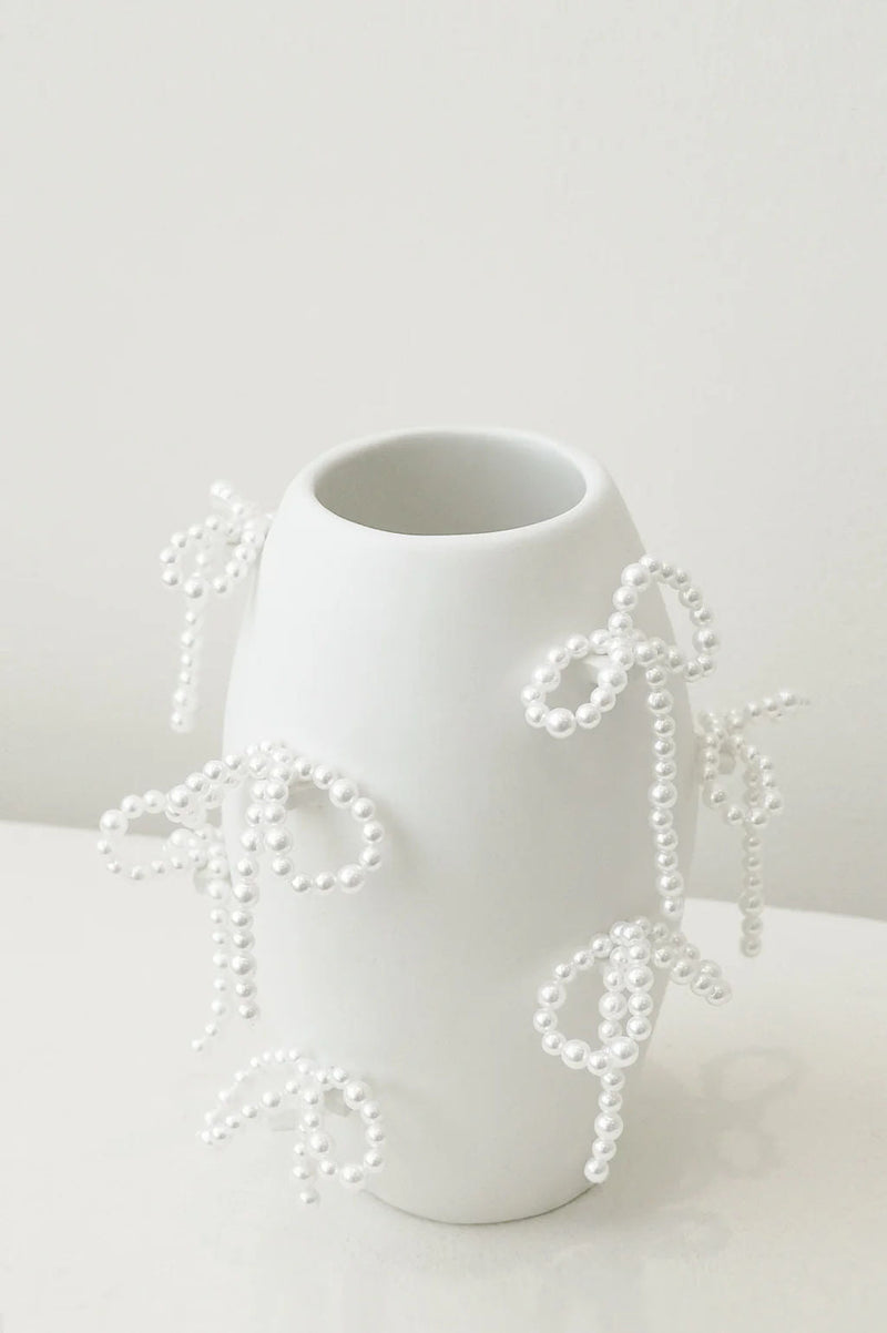 The "Notsobig" Power, Corruption, Lies Small Vase in matte white by Completedworks features faux pearl bows and sits elegantly on a white surface against a light background.