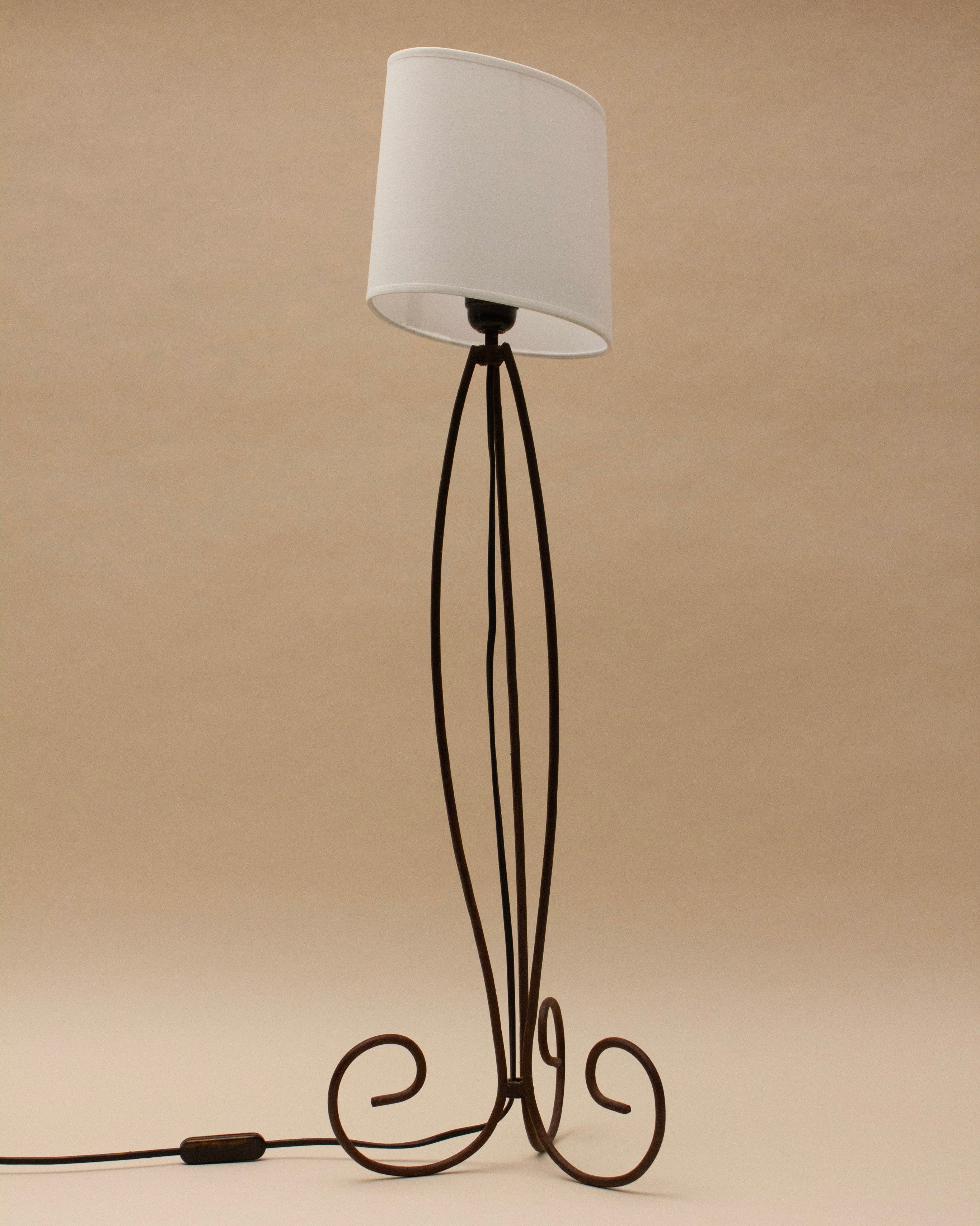 The SÒLER SANTA CRUZ DE TENERIF LAMP features a tall, decorative brown wrought iron stand with curled feet and a white cylindrical shade. The power cord and switch are visible on the left against a plain beige background.