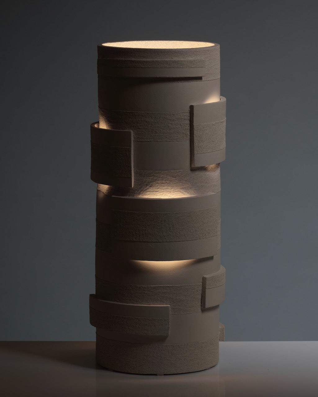 The "Bricks" Table Lamp in ceramic by Katia Langeard features textured, layered surfaces with rectangular accents, casting soft light through its gaps—an elegant accent for a hallway atop a reflective surface against a plain background.