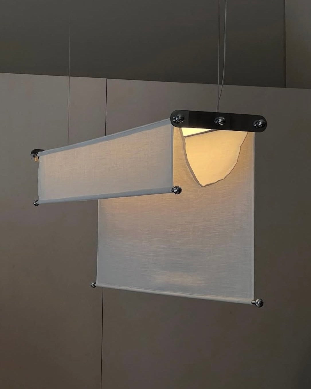 The Vento Pendant by Caia Leifsdotter is a modern adjustable light with two layered rectangular fabric panels, the upper partly torn to reveal a glowing bulb. Suspended by thin wires, it adds an organic touch to any space.