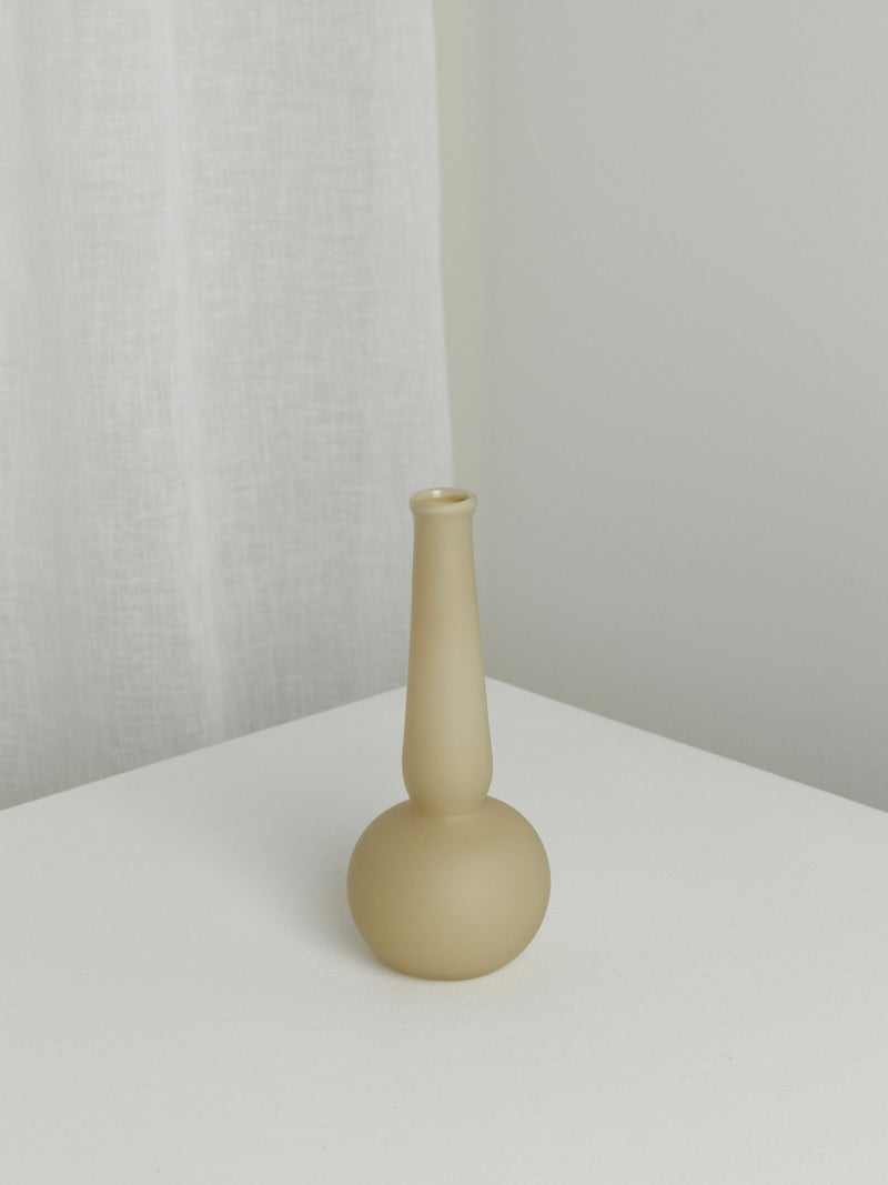 The Verre d'Onge Small Vase in Caramel, featuring a matte beige ceramic finish with a narrow neck and round base, sits on a white surface by a white curtain and light gray wall.