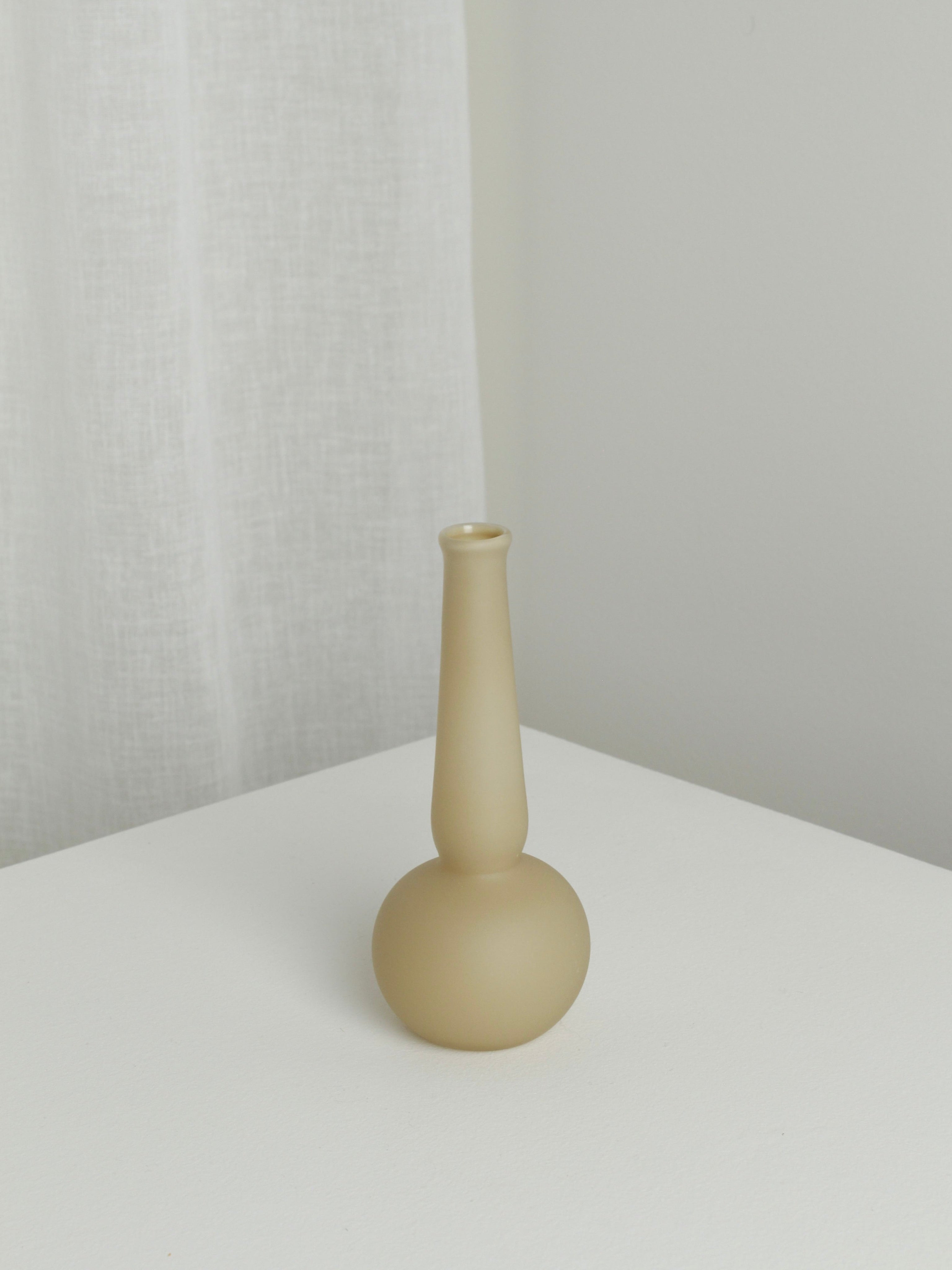 The Verre d'Onge Small Vase in Caramel, featuring a matte beige ceramic finish with a narrow neck and round base, sits on a white surface by a white curtain and light gray wall.