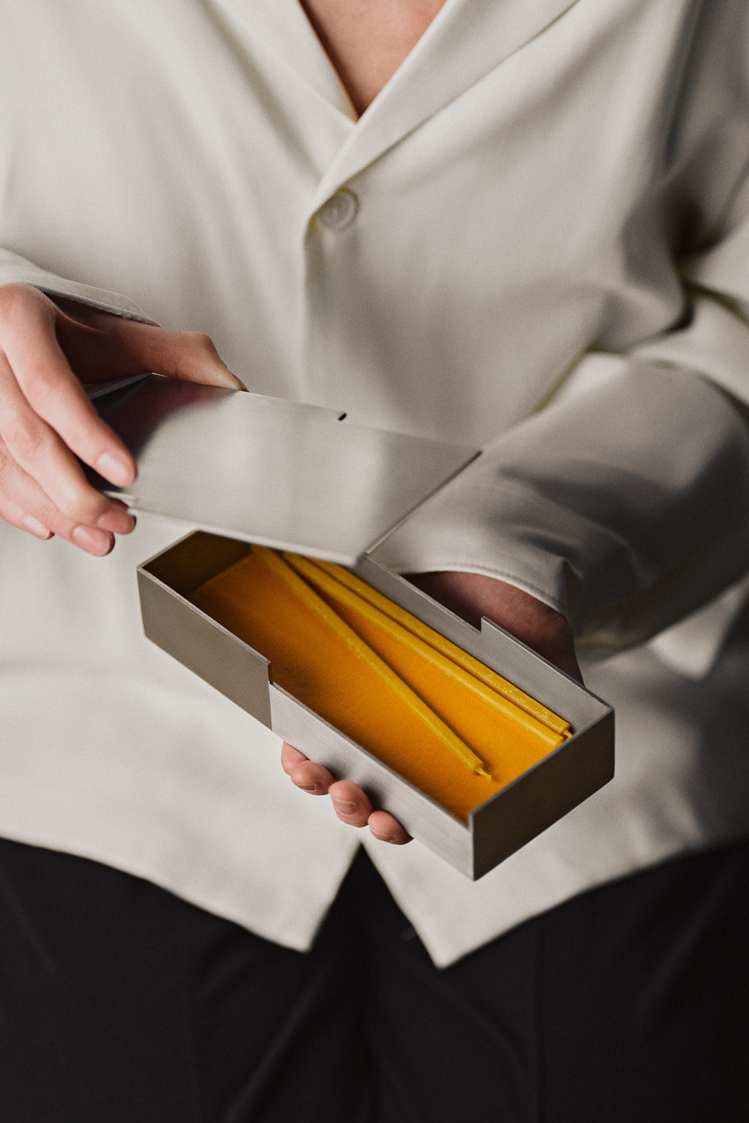 A person in a cream-colored shirt holds the open HYZ Carlo - Medium desk set, a handcrafted steel box lined with yellow fabric, revealing a yellow pencil inside—a unique desk accessory.