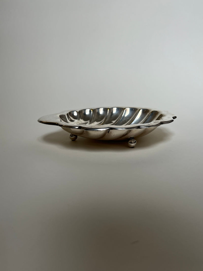 The Manushi Medium Shell Shaped Vide Poche features a shallow, oval form with a scalloped design and rests on three small spherical feet, shown against a plain light background.