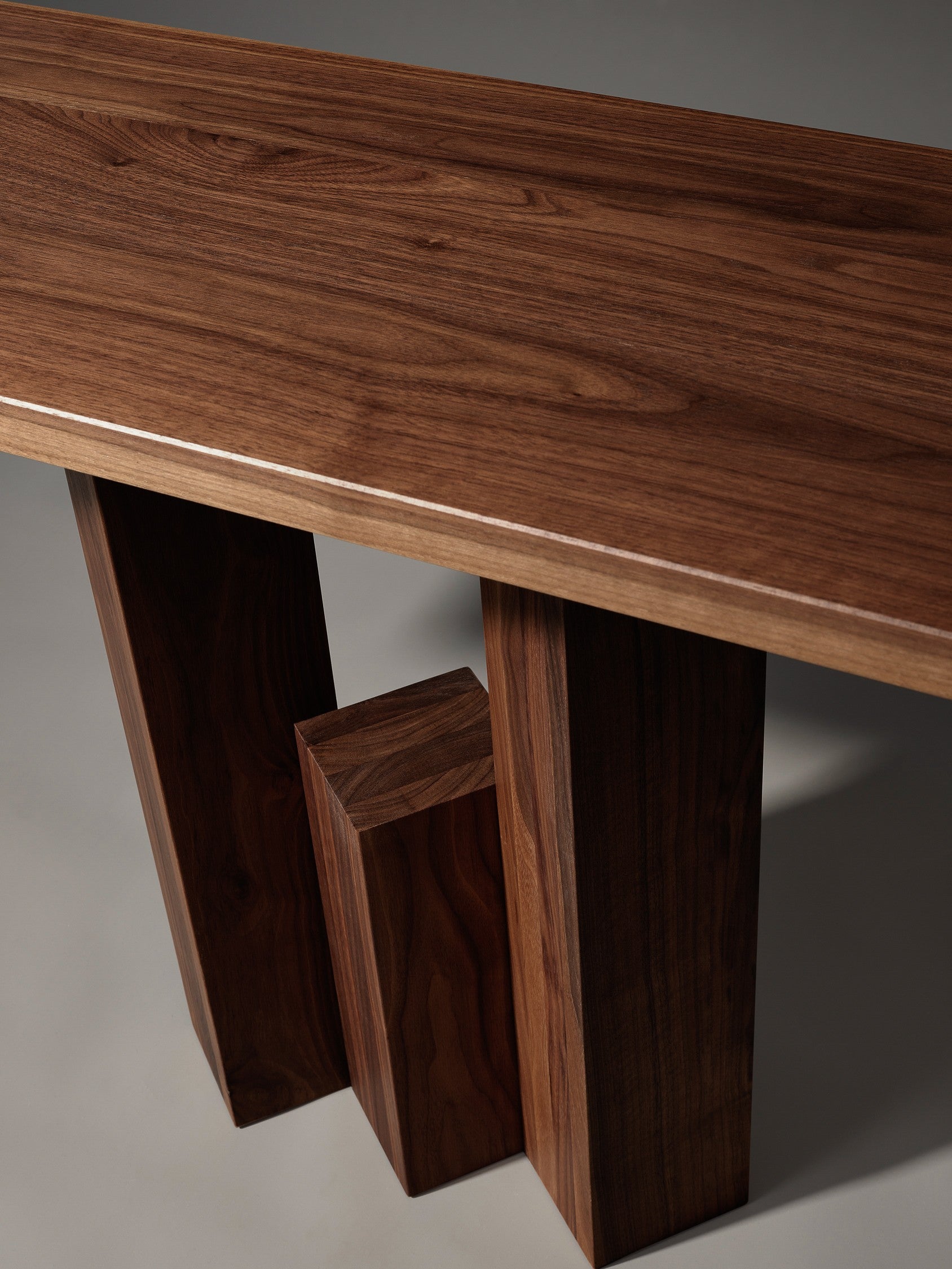 The Fenestra Console by Mokko features a smooth, polished dark wood surface with visible grain and three thick, rectangular legs of varying heights. This elegant, customizable piece brings individuality to any space.