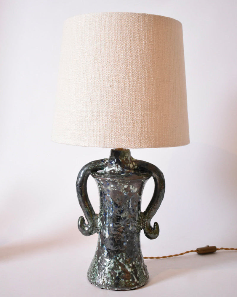 The "Kasaï" Erato Blue and Green Ceramic Table Lamp by Barracuda Interiors features a textured base with two curved handles and a light beige fabric shade, plugged in and set against a plain white background.