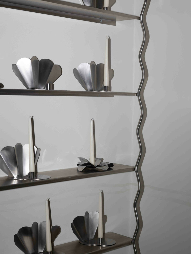 The Good Living&Co. Wave Shelves feature a wavy metal frame on the right, displaying modern, flower-shaped candle holders with white tapered candles against a plain, light background.