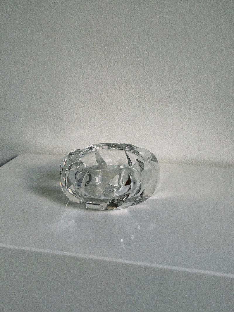 The Manushi Saint-Louis Crystal Ashtray, with its clear textured design, rests on a glossy white surface against a light gray wall. The minimal, softly lit scene highlights the ashtray's luxurious reflections and transparency.