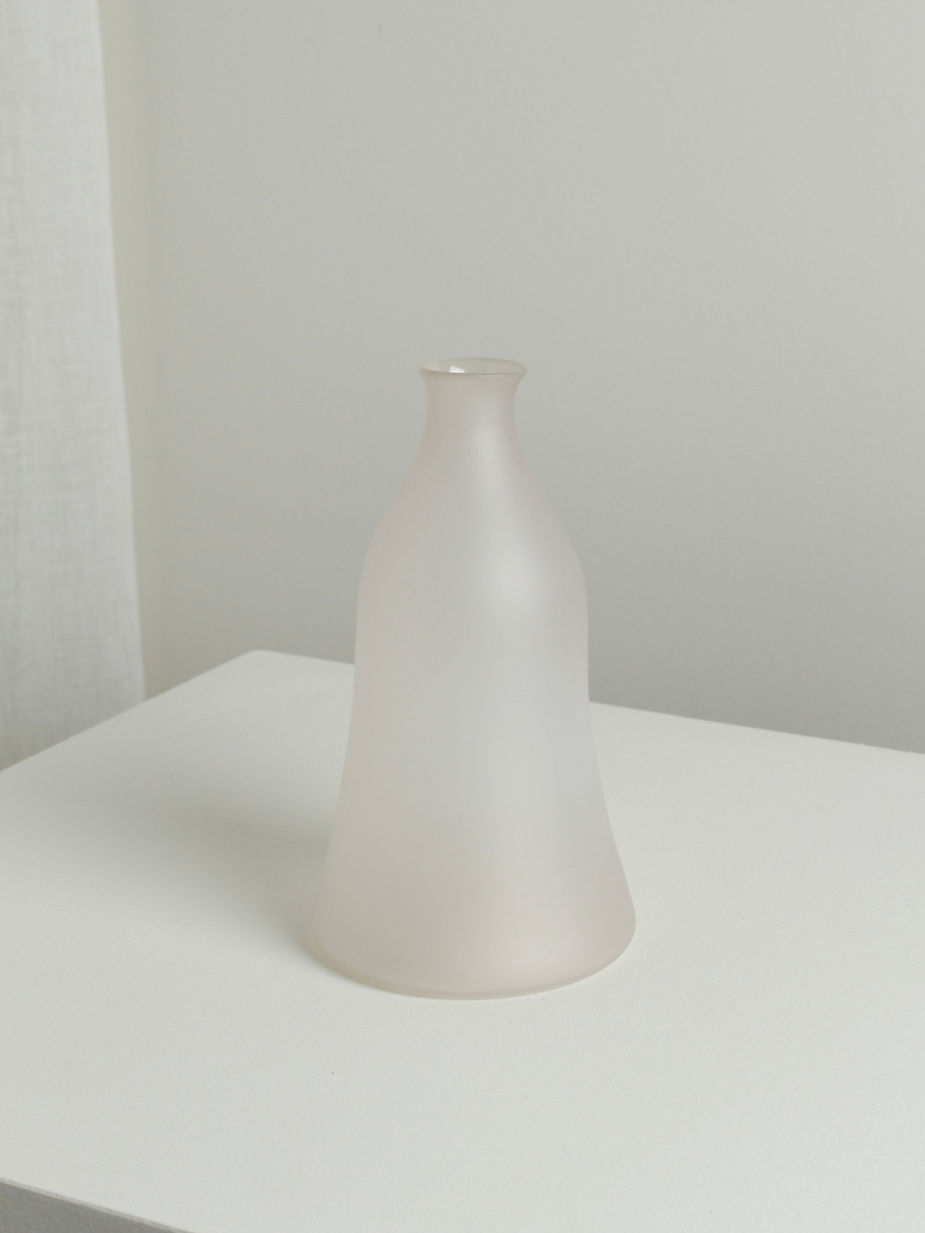 The Medium Vase in Moon by Verre d'Onge, featuring frosted glass with a narrow neck and wide base, rests on a white surface against a light-colored wall in a minimalist setting.