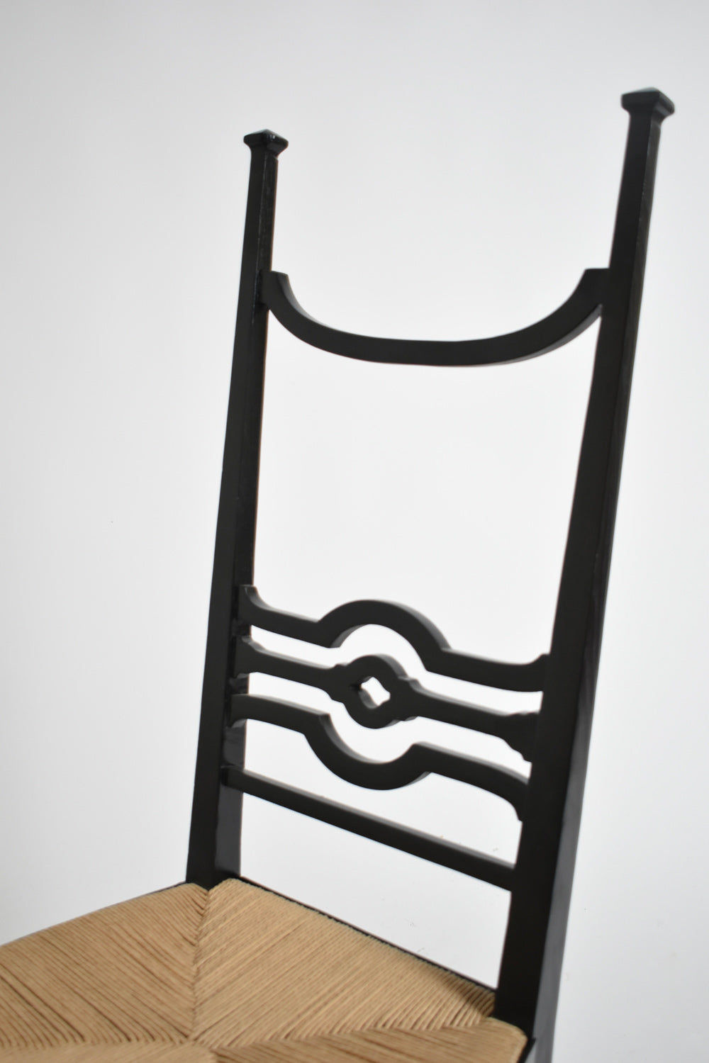 A close-up of Barracuda Interiors’ Arts & Crafts Ebonised Wood Chair, 1900s, with a tall geometric cut-out backrest and a natural woven seat, set against a plain white background.