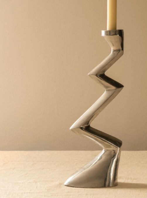 The Les Objets Amsterdam Matthew Hilton Zigzag Candle Holder, featuring a modern silver design, displays a single light-colored candle atop a neutral textured surface with a beige background.