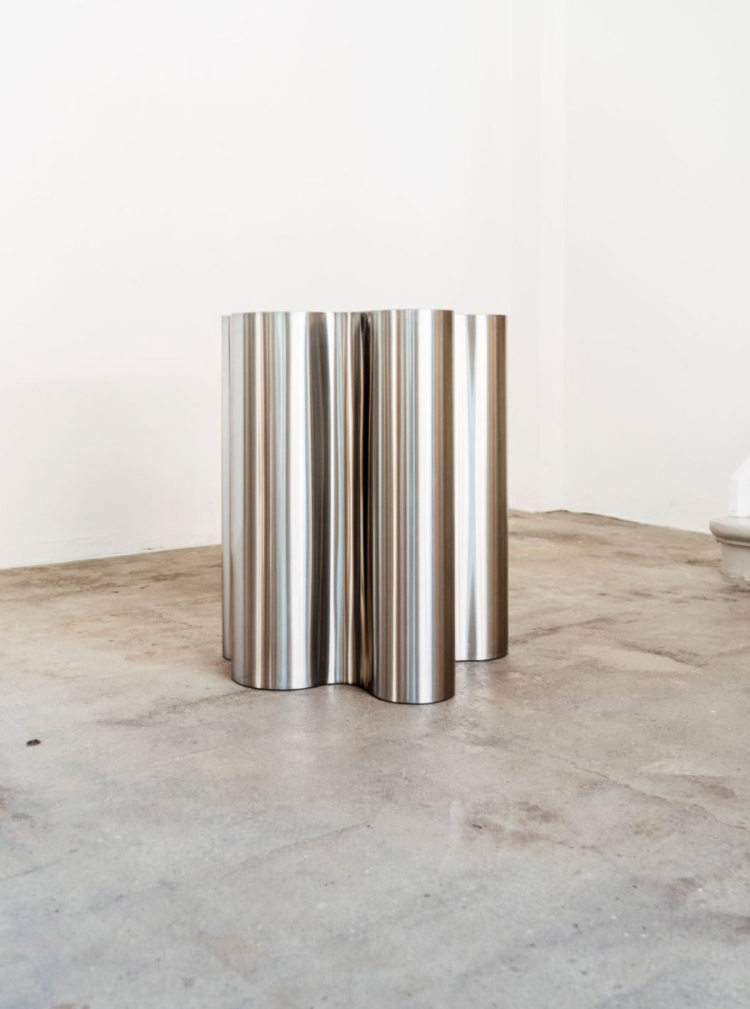 The Silver Root - Tall side table by Caia Leifsdotter features a sculptural design with four connected cylindrical steel forms and a polished, reflective finish, shown on a concrete floor in a minimalist white-walled room.