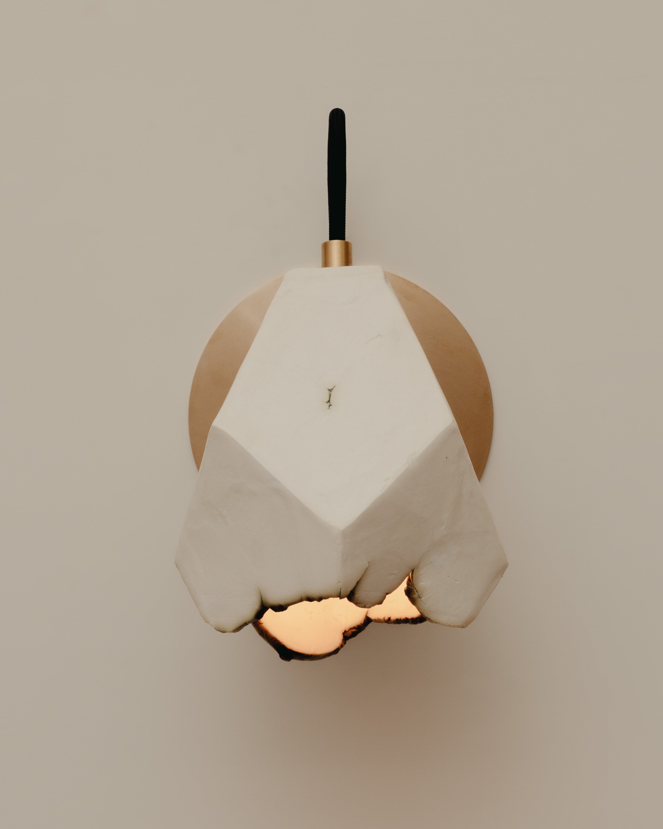 The Relic Spotlight Sconce by JS/WRKSHP features a geometric white sculpted shade and gold circular base, casting warm light against a beige wall for elegant, modern lighting.