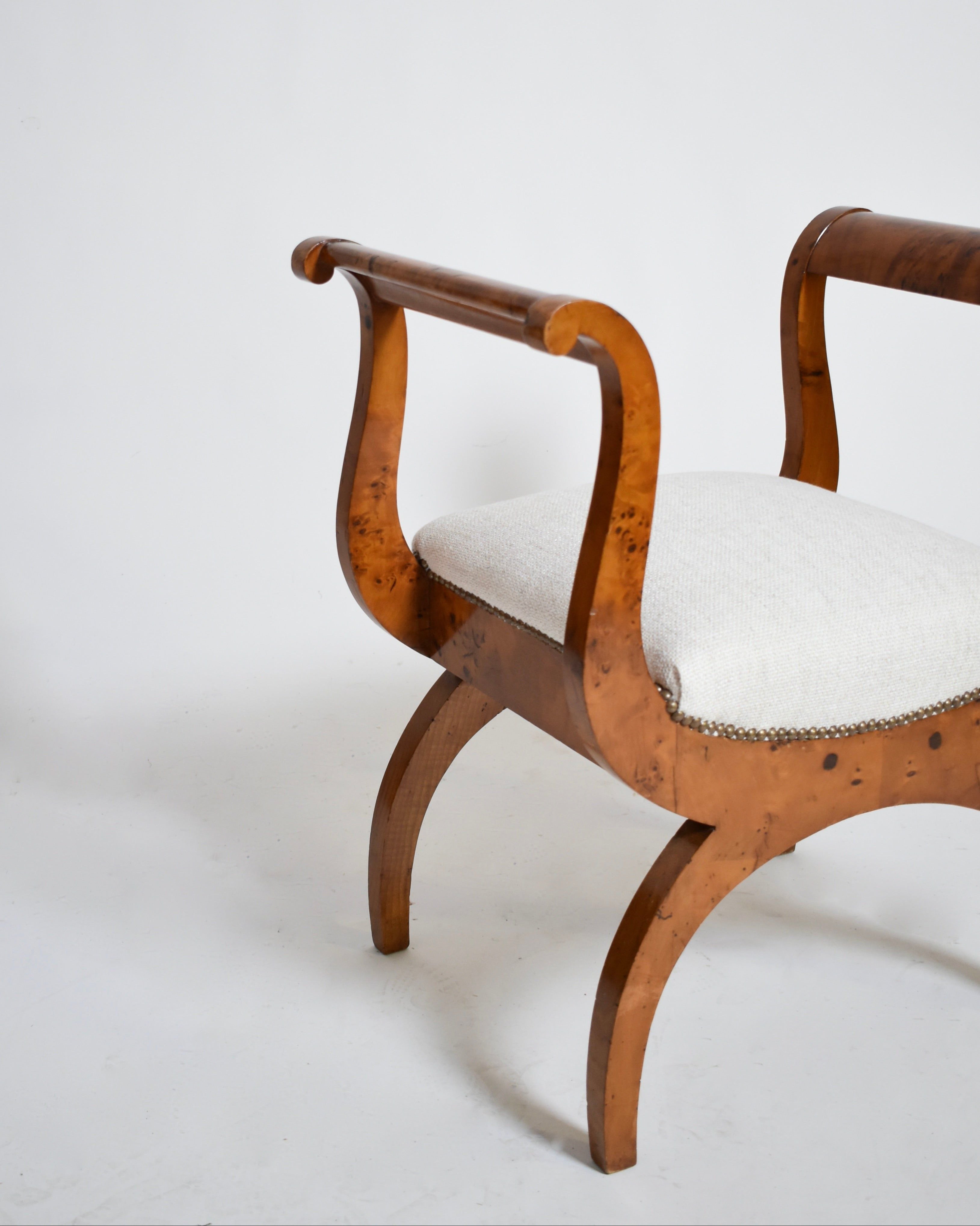 A pair of Walnut Curule Stools, XIXth by Barracuda Interiors, with curved armrests and legs and a light upholstered seat with nailhead trim, set against a plain white background.
