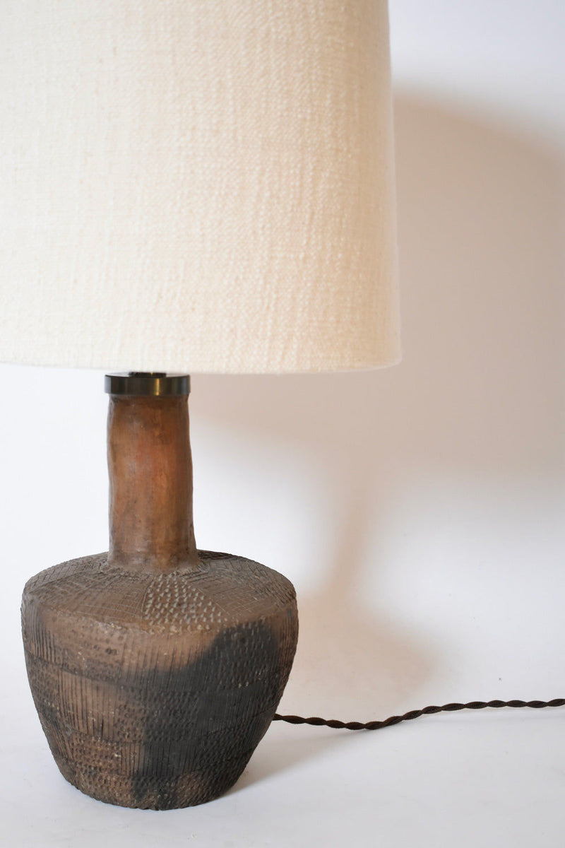 The African Brown Ceramic Table Lamp by Barracuda Interiors, 1970s, features a textured dark brown terracotta base with a subtle geometric pattern and a large off-white fabric shade. The twisted cord extends to the right on a plain white background.