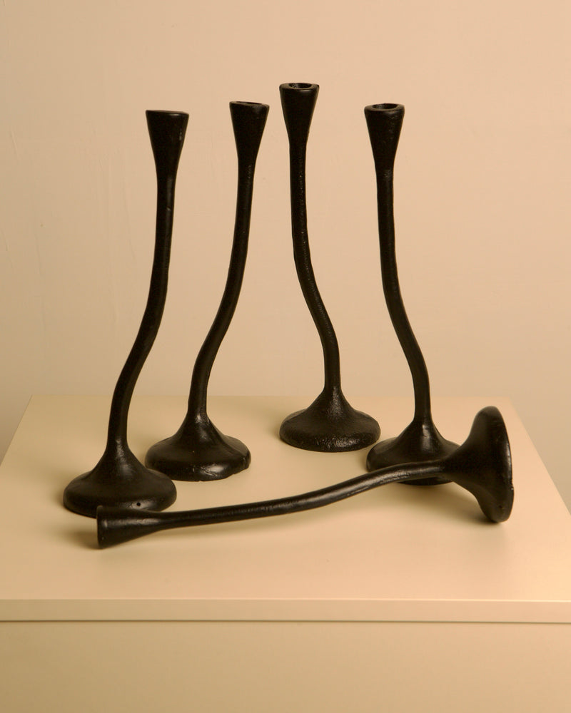 A set of 5 "Twisted" Candleholders in black cast aluminum by Treaptyque, inspired by 80s design, are arranged on a white pedestal—four upright and one horizontal—against a plain light-colored wall.