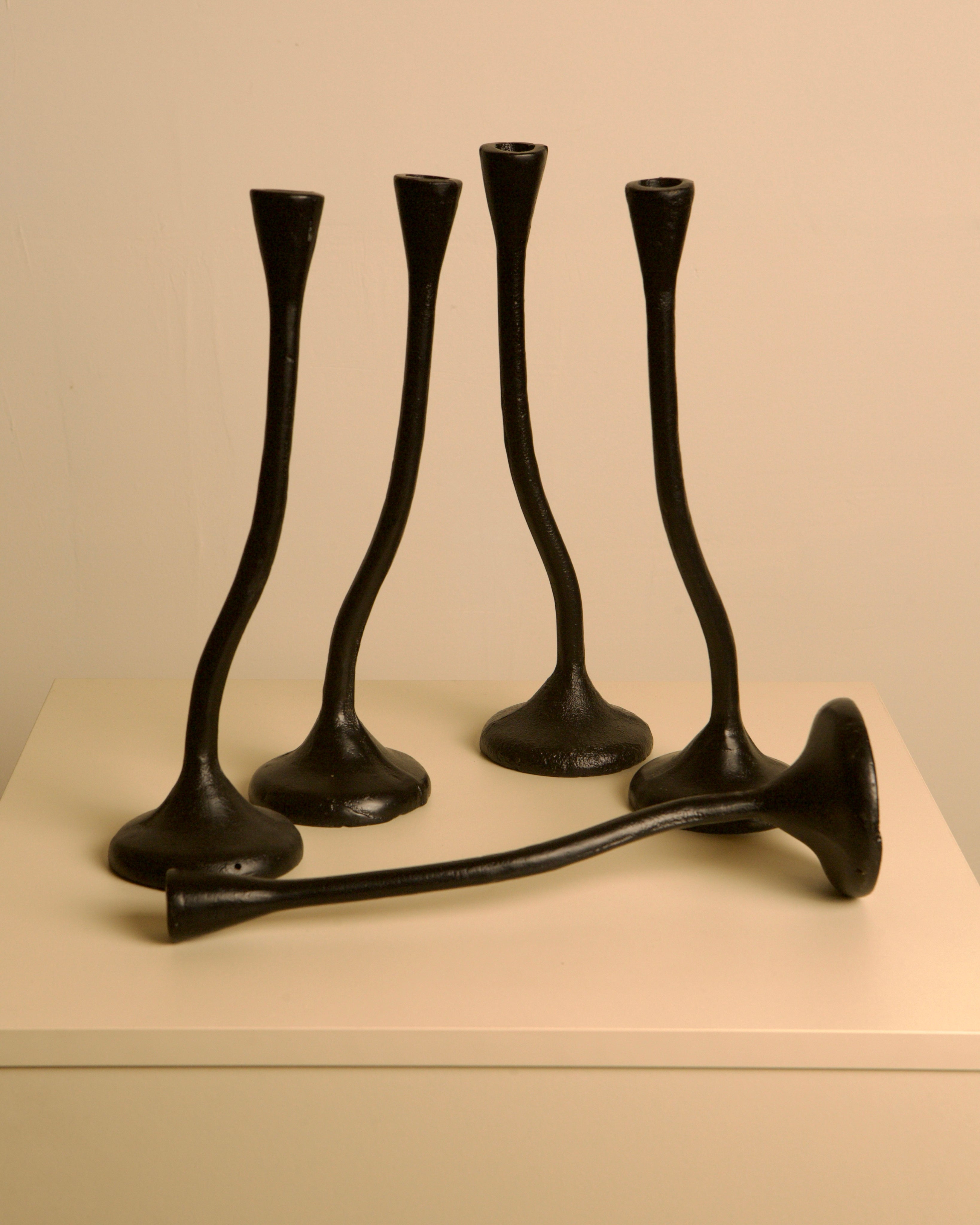 A set of 5 "Twisted" Candleholders in black cast aluminum by Treaptyque, inspired by 80s design, are arranged on a white pedestal—four upright and one horizontal—against a plain light-colored wall.
