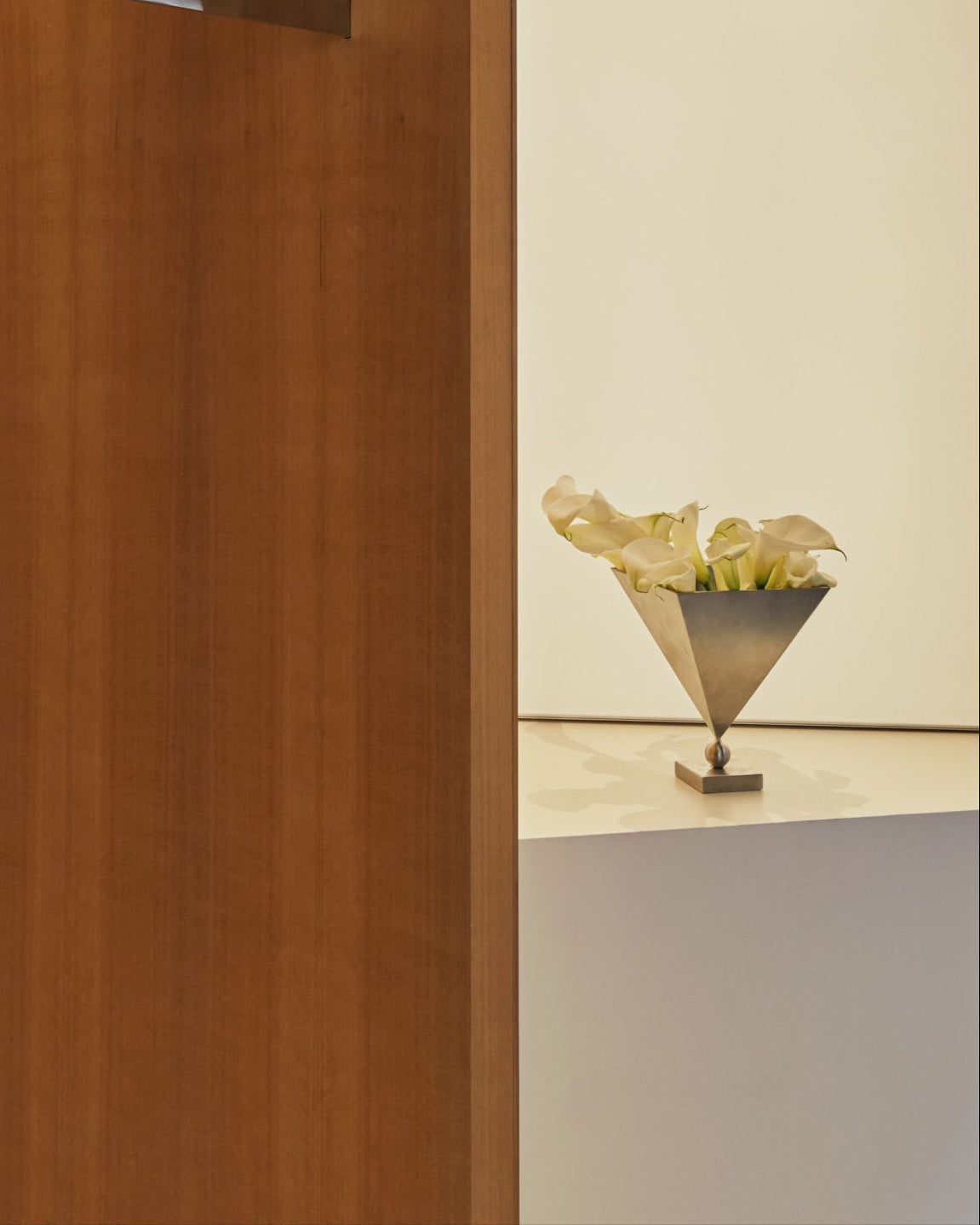 The Stainless Steel 'Vasen' Vase by Studio Vraco, shaped in a modern triangle and holding white calla lilies, rests on a white surface and is partially hidden by a vertical wooden panel in a minimalist setting.