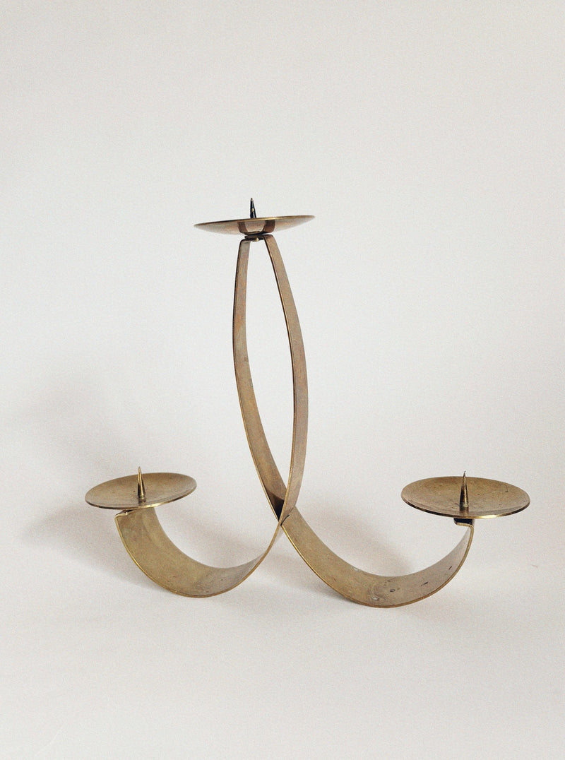 The Huerto 2 Curved Brass Candleholders feature elegant curved arms and round plates with central spikes, blending modern style with vintage charm against a plain white backdrop.