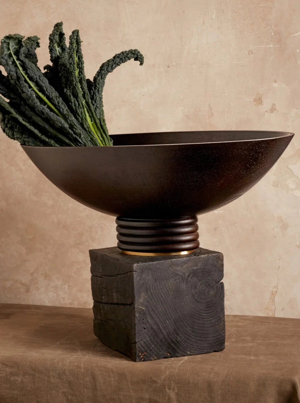 A large Alhambra Oval Bowl by LOBJET resting on a square, textured brass base. The bowl contains tall, leafy green vegetables (kale) extending upward. It's placed against a neutral beige backdrop.