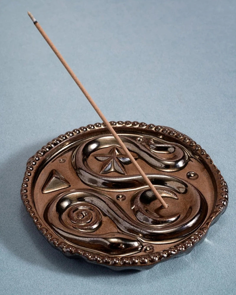 A detailed, round metal incense holder with intricate patterns, holding a single burning incense stick at an angle. The holder sits on a flat, light blue surface, and the designs on the holder include swirls and geometric shapes reminiscent of Greek mythology. The Lobjet Earthenware Incense Holder is perfect for adding a touch of elegance to your home fragrance collection.