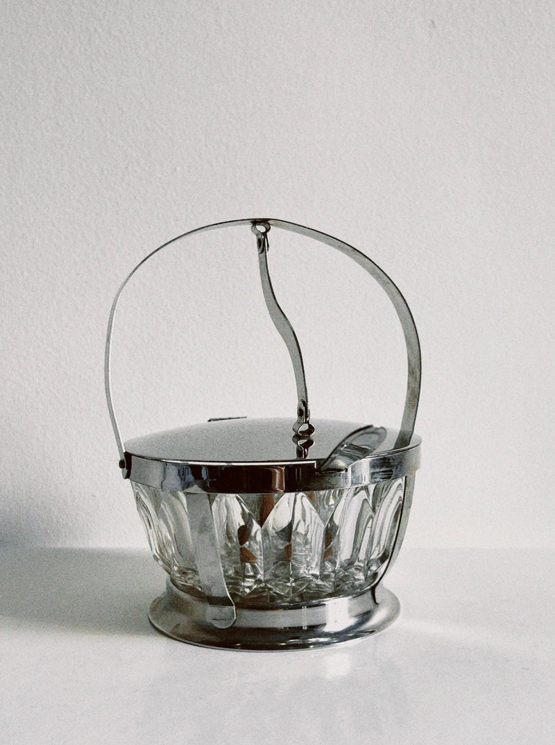 The Manushi Crystal Sugar Bowl, featuring a metal lid and curved handle, serves as an elegant ice bucket. Shown on a white surface with a plain light background.