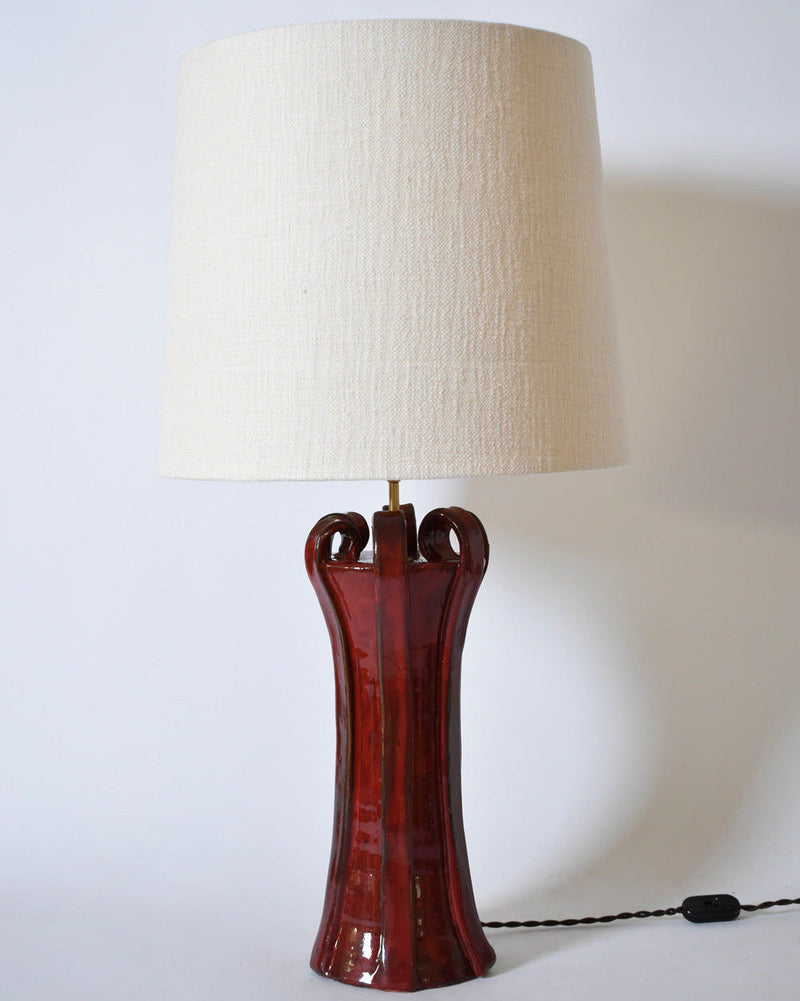 The "Kasaï" Henry Red Ceramic Lamp by Barracuda Interiors features a glossy Albarelo-inspired base with curved top accents and a large cylindrical off-white fabric shade. The lamp is shown plugged in, with its cord visible on the right.