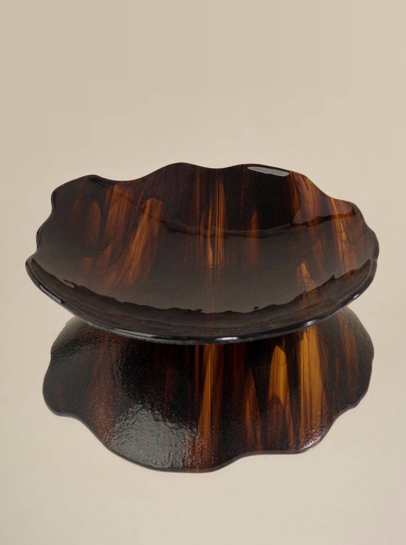 A dark, wooden, wavy-edged Plate Carey Fig by Los Objetos Decorativos displayed on a reflective surface, creating a clear shadow below it. The plate's rich brown tones have swirling patterns characteristic of artisanal manufacturing.