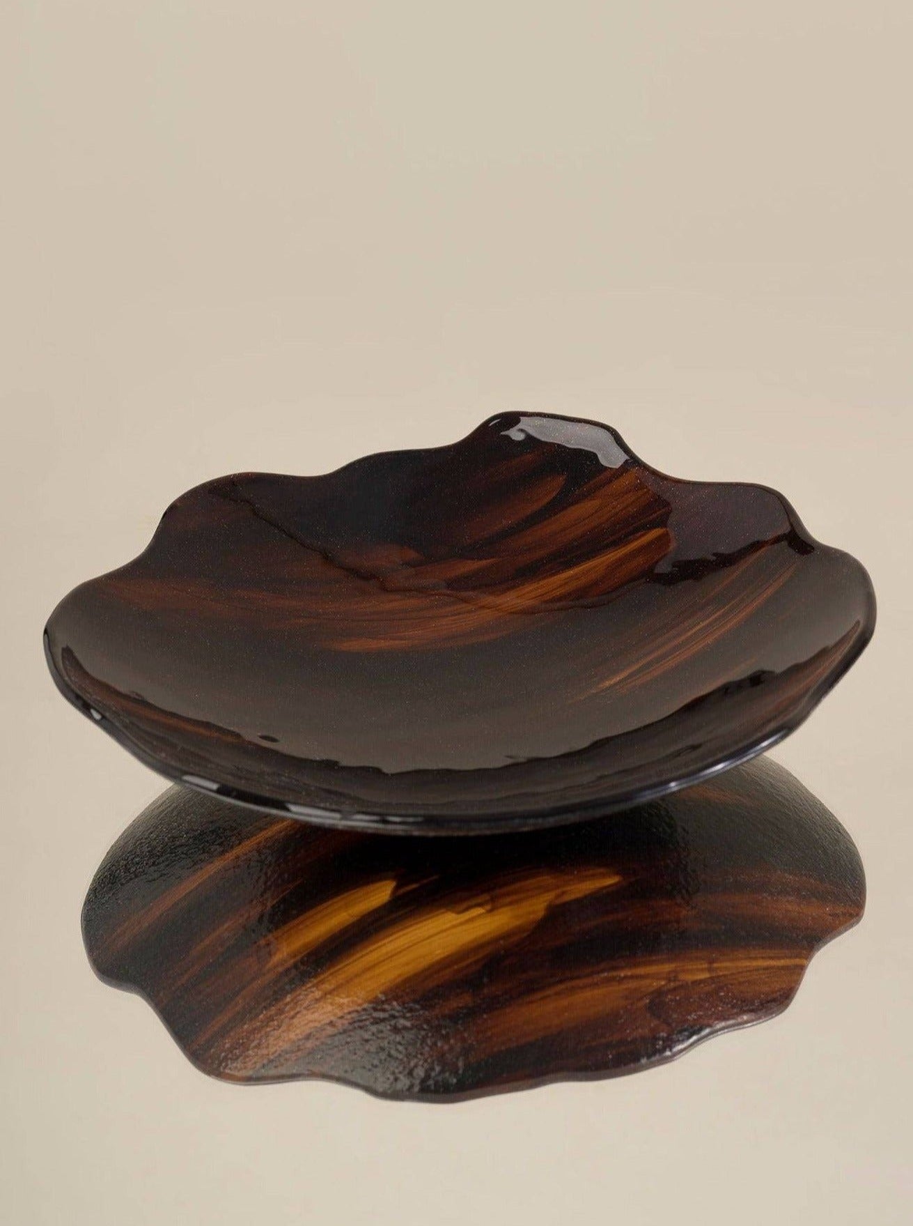 A dark-brown, swirling, wood-grain patterned Plate Carey Fig vessel on a light beige background, casting a reflective shadow from Los Objetos Decorativos.