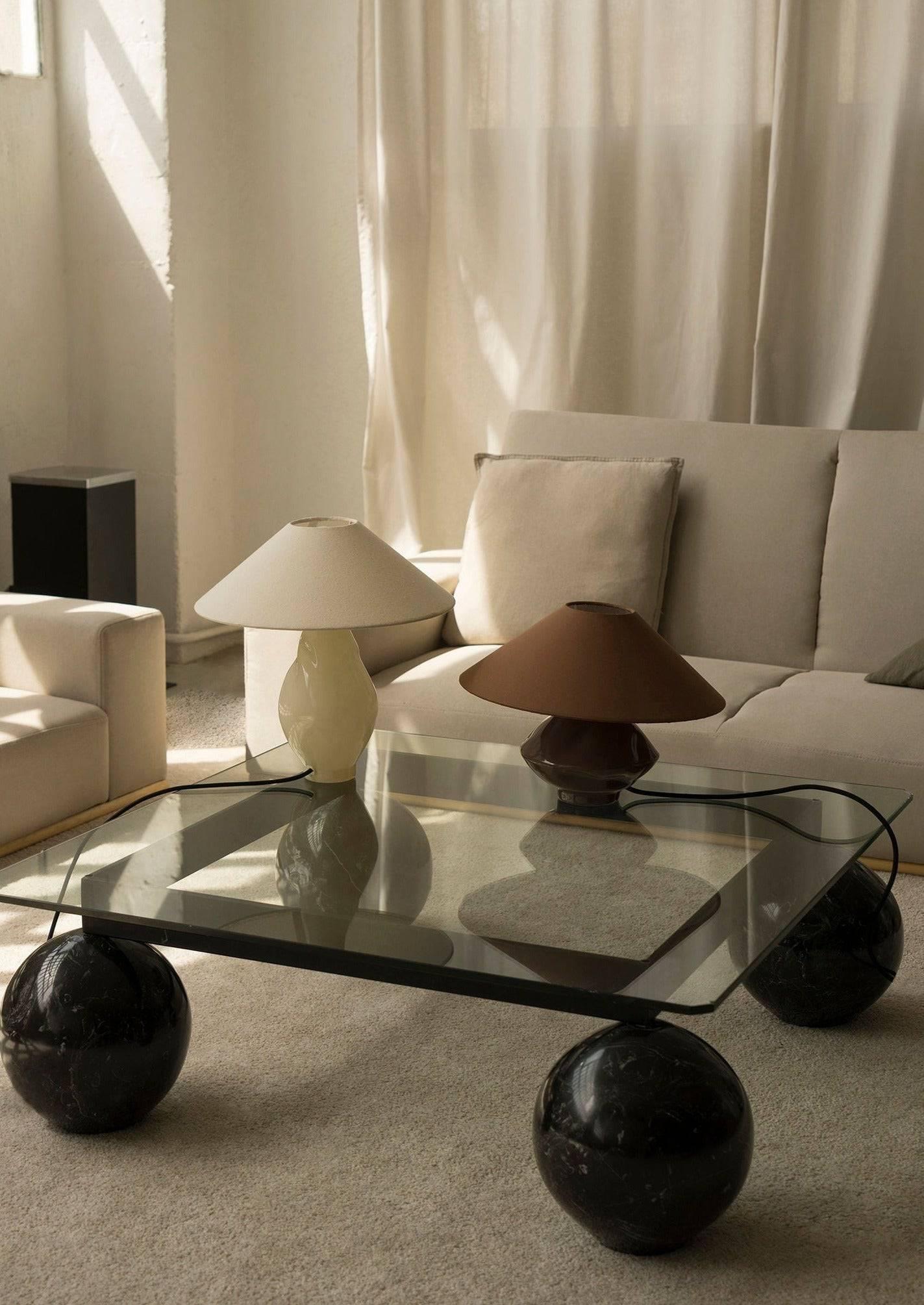 A modern living room featuring a glass coffee table supported by black spherical legs, two Los Objetos Decorativos beige conical glass lamps, and a beige sofa in a room with soft natural lighting and white curtains exuding interior elegance.