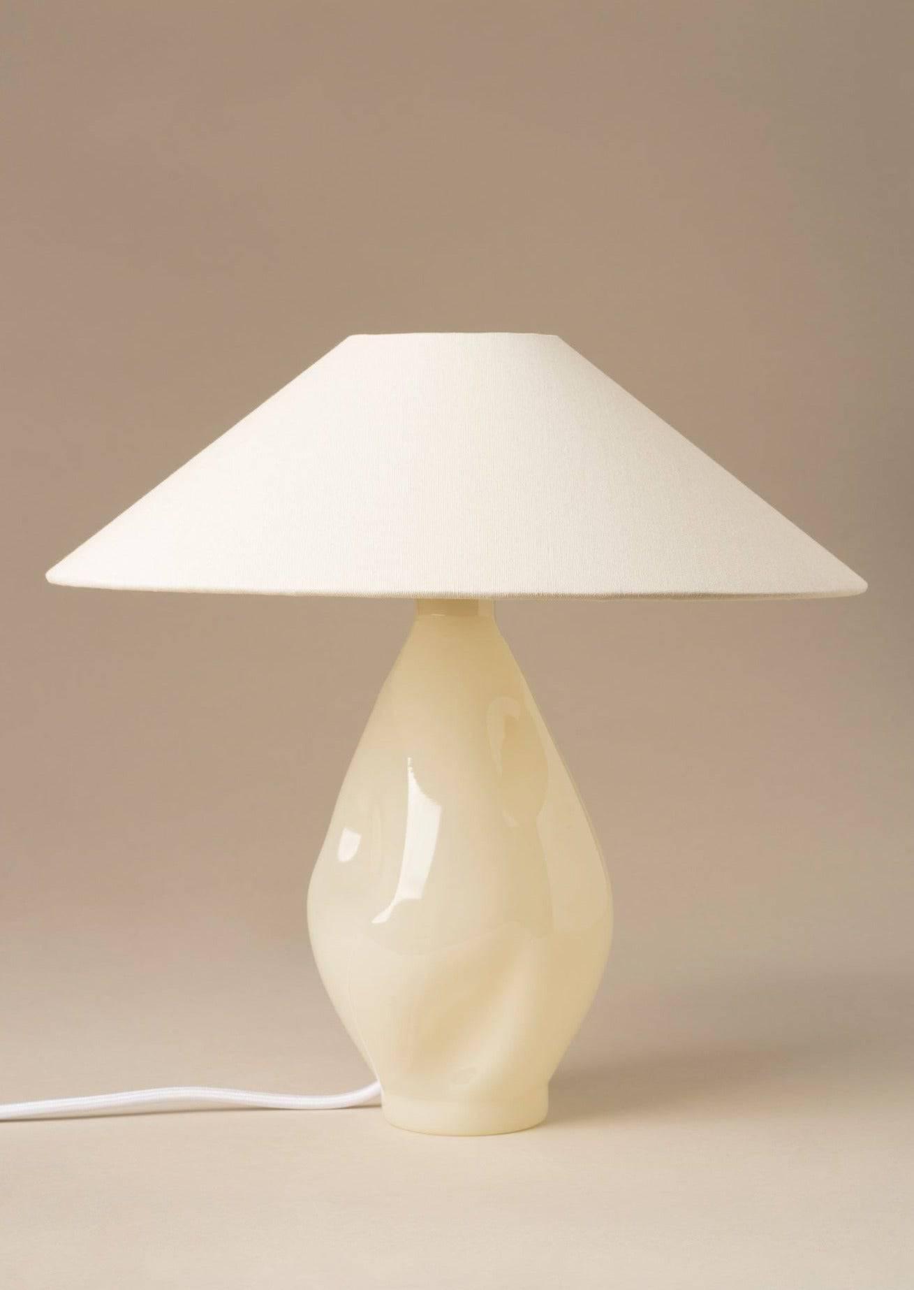 A cream-colored table lamp with a glossy ceramic base and a large, flat, Beige Conical Glass shade, set against a neutral beige background by Los Objetos Decorativos.