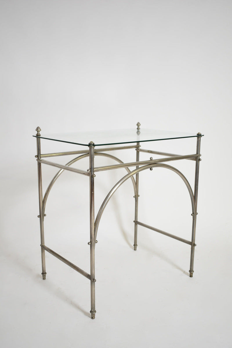The Tubular Chrome Iron and Glass Table by Barracuda Interiors features a clear glass top, chrome iron frame with arched supports and finials, capturing 1920s style. Shown against a plain white background.