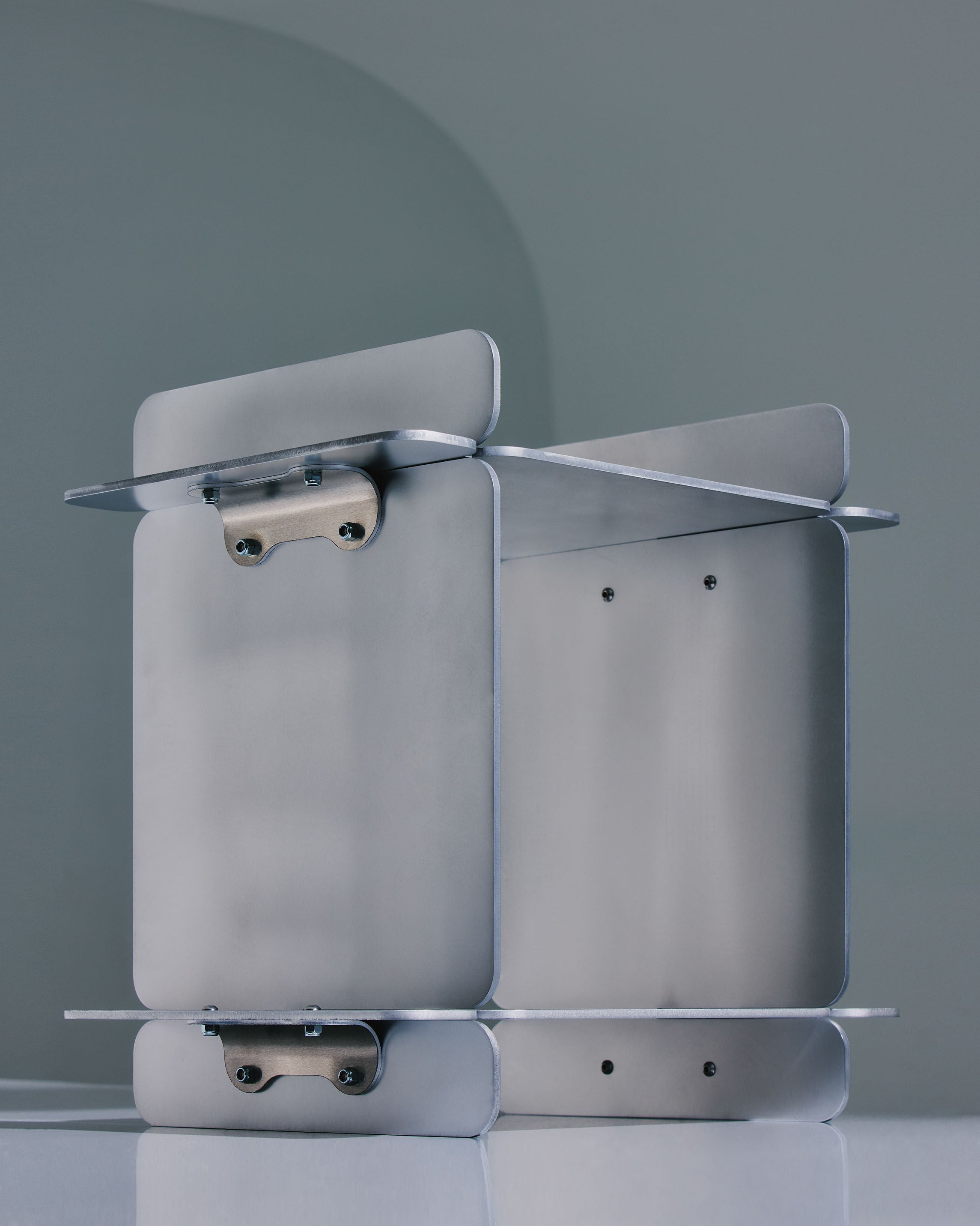 The Side Table by LESORR is a minimalist piece with a metallic frame, flat rectangular panels, rounded edges, and visible bolts and hinges, shown against a muted blue-grey background.