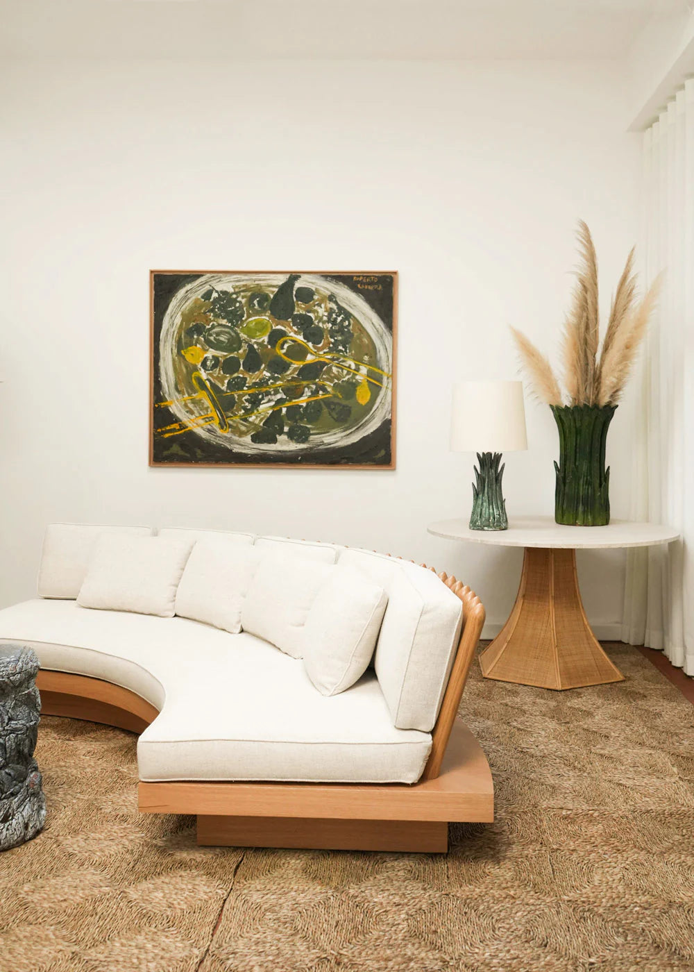 A modern living room featuring the San Romano Round Oak Sofa by Barracuda Interiors, a round wooden table with a lamp, tall pampas grass, textured carpet, and an abstract green, yellow, and white painting on a crisp white wall.