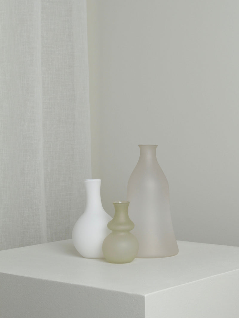 The Verre d'Onge Medium Vase in Moon, with its minimalist frosted glass design, is arranged on a white pedestal against a neutral background with a sheer curtain to the left.