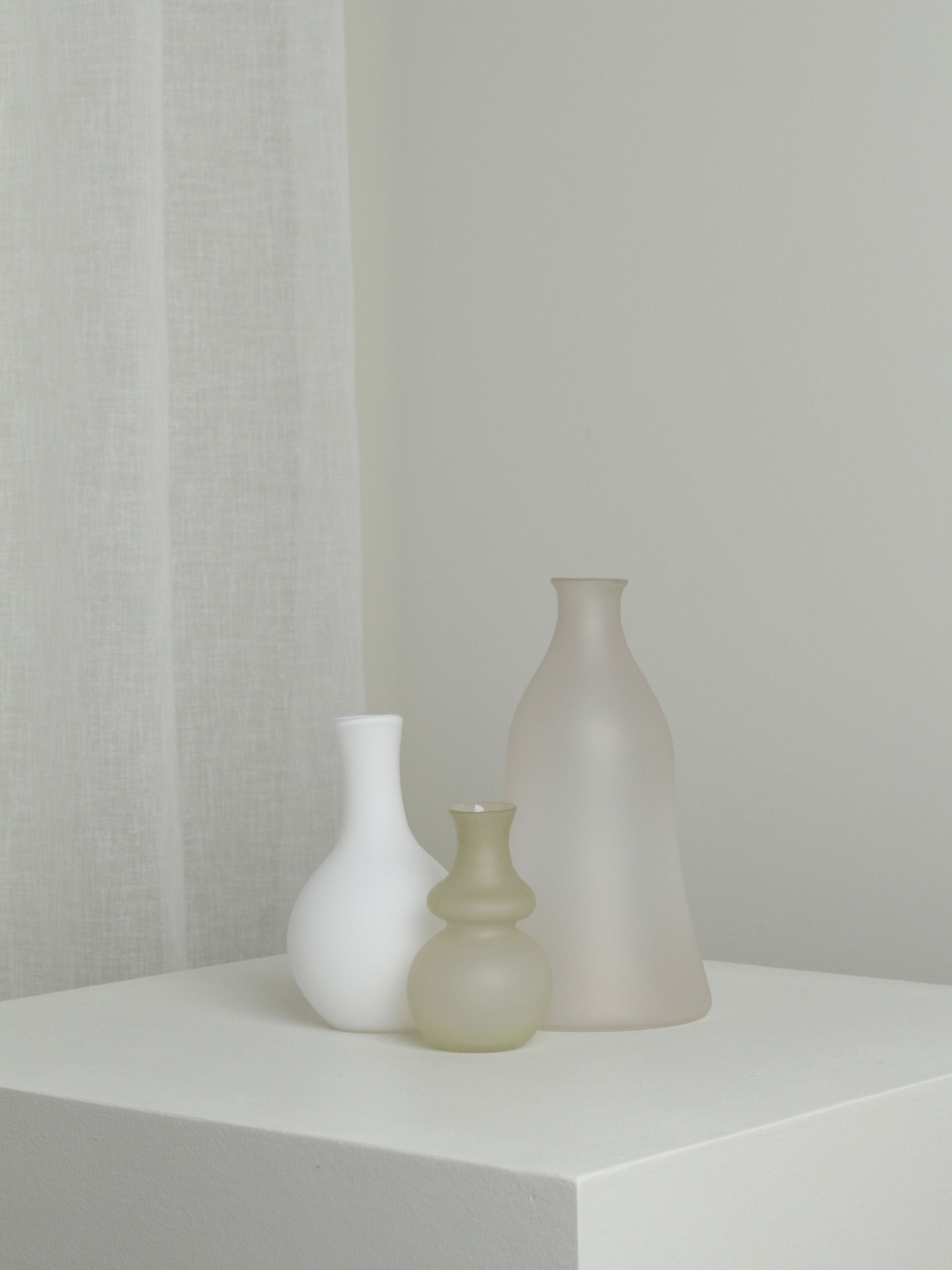 The Verre d'Onge Medium Vase in Moon, with its minimalist frosted glass design, is arranged on a white pedestal against a neutral background with a sheer curtain to the left.