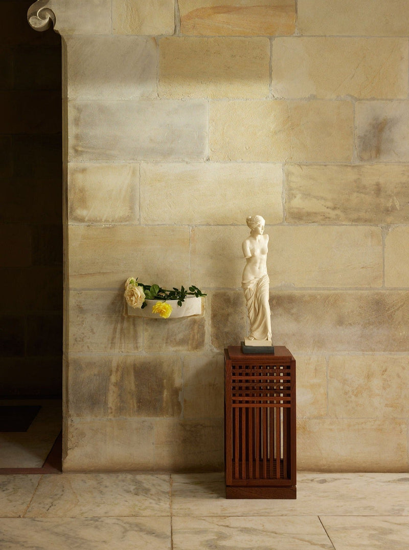Riviera Plinth short in Oiled African Mahogany — positioned beside a stone wall