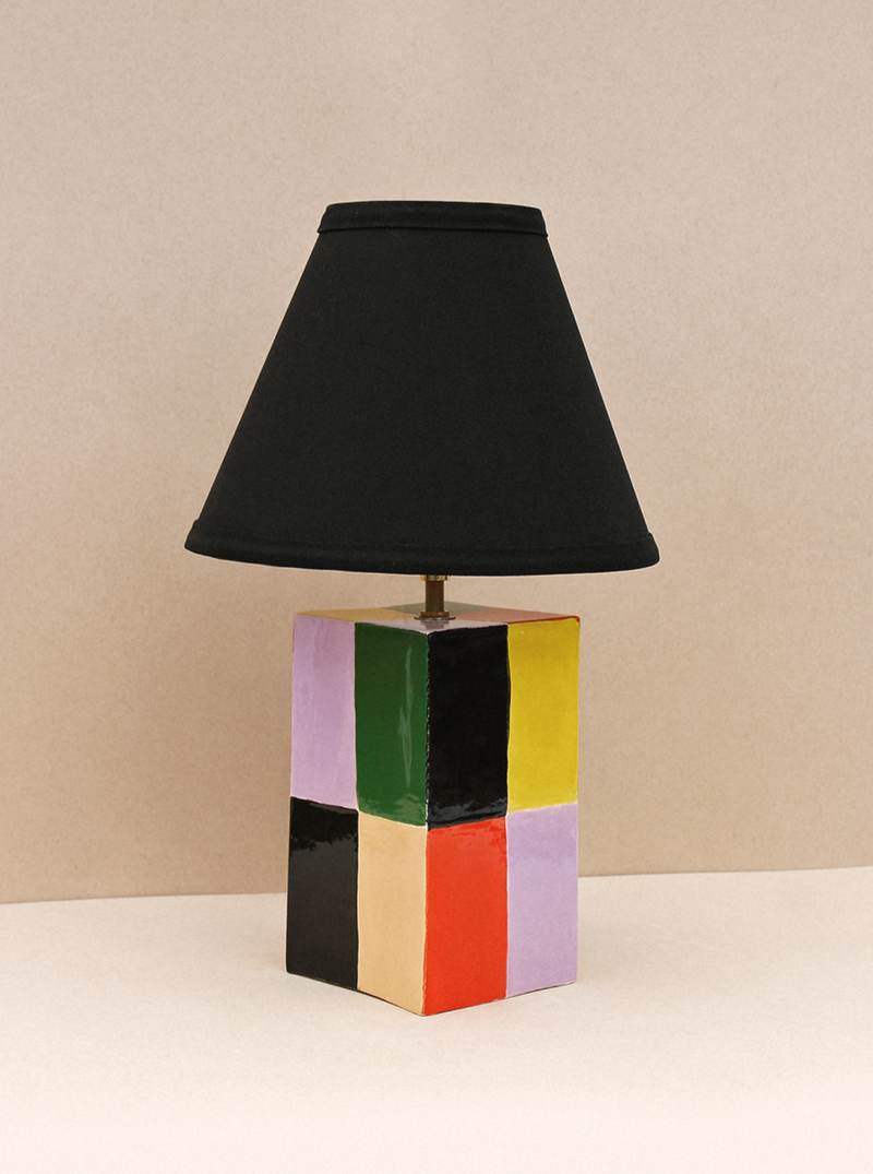 A colorful, cubic Milagro Lamp (Chico) with a black shade, set against a neutral-toned background. The base features bright, multicolored panels in green, yellow, red, purple, and black by Casa Veronica.