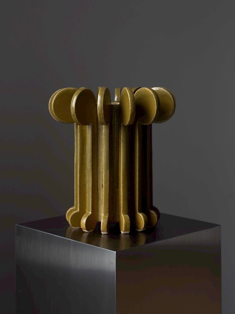 Vessel No. IV by Devin Wilde is a golden abstract sculpture of vertical, rounded, interlocking forms on a dark reflective pedestal, set against a plain gray background.