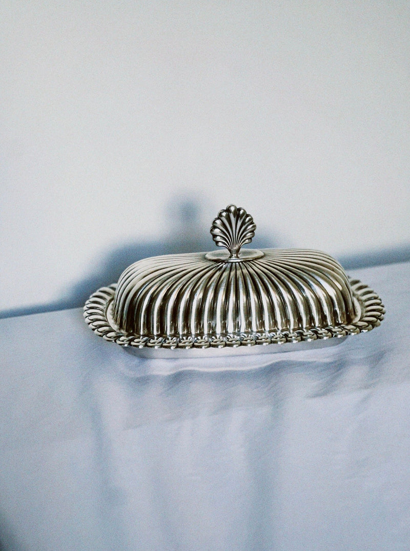 The Manushi Silver Shell Detailed Butter Dish with a ribbed lid and decorative shell-shaped handle rests on a white tablecloth against a plain, light background.