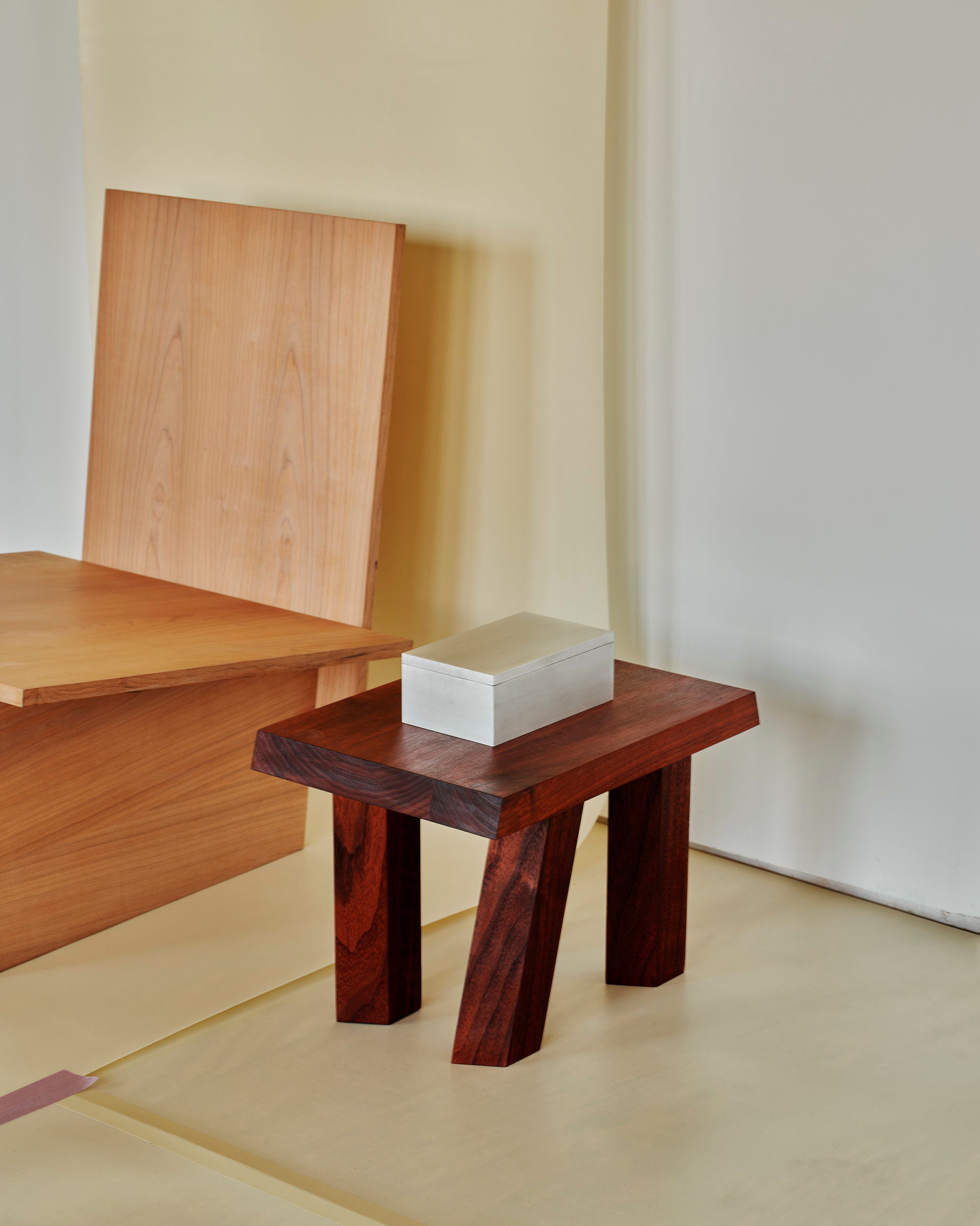 A minimalist setup featuring a wooden bench with a simple backrest made of vertical wood panels. Next to it is a small, two-legged wooden stool with an aluminum MOCK STUDIO Miter Box placed on top. The background is neutral, emphasizing the clean lines and natural wood texture.