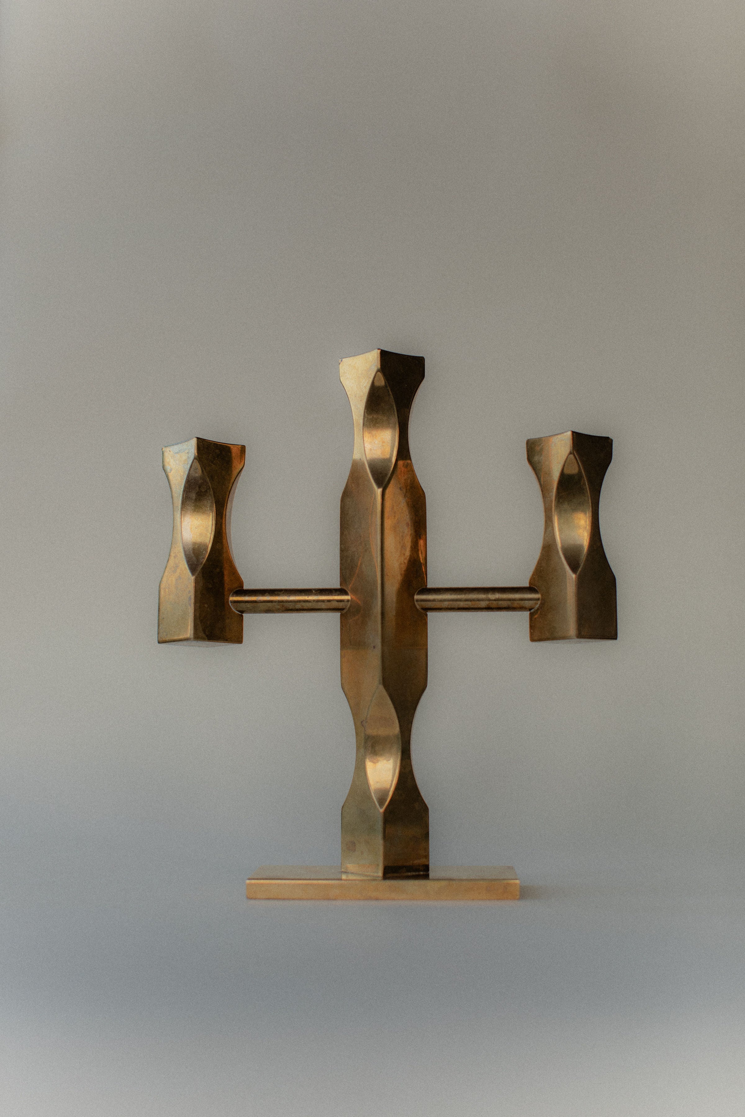 Out For Lunch's Brass Candleholder is a modern brass and wood sculpture with a central vertical piece and two symmetrical, angular arms extending horizontally. Reminiscent of Brutalist design, it rests on a flat wooden base against a neutral background.