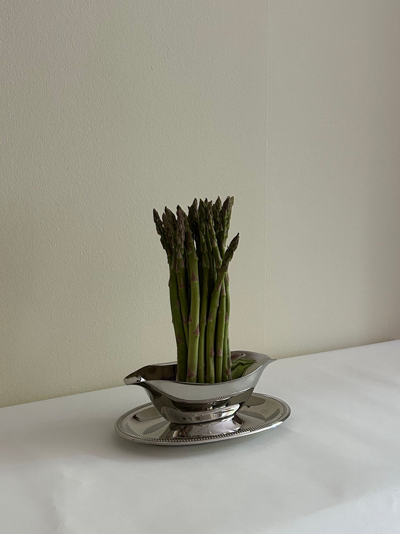 A bundle of asparagus stalks is arranged vertically in a Flock Studio Stainless Steel Server Sauce on a matching tray, set on a white dining table against a light-colored wall. The polished finish enhances the minimalist and elegant aesthetic of the scene.