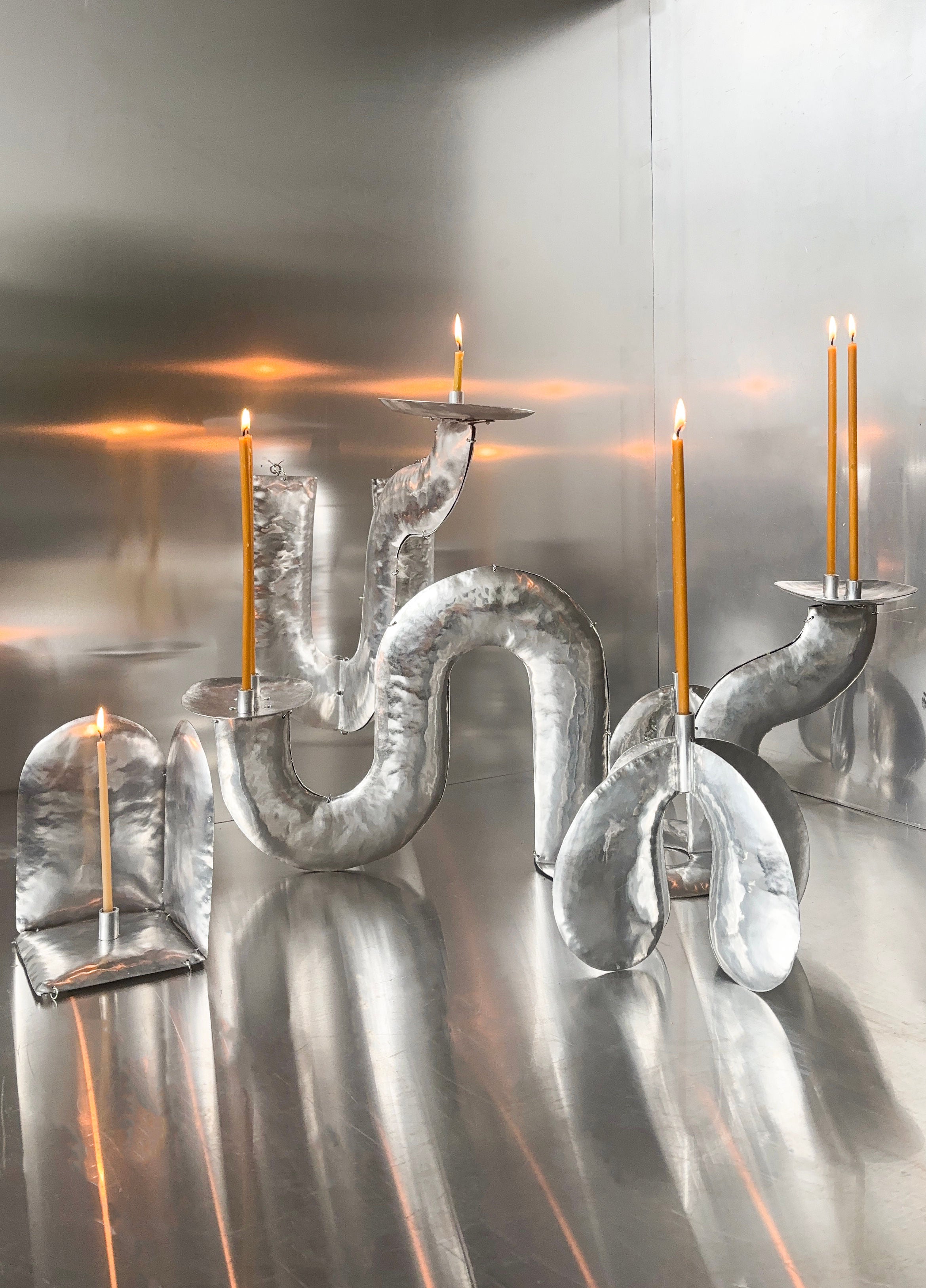 Four beeswax candles adorn the Phaedra candle holder LARGE by Bregje Sliepenbeek, crafted in Amsterdam. These silver candlesticks boast flowing, wavy designs against a metallic backdrop, offering a modern and artistic vibe.