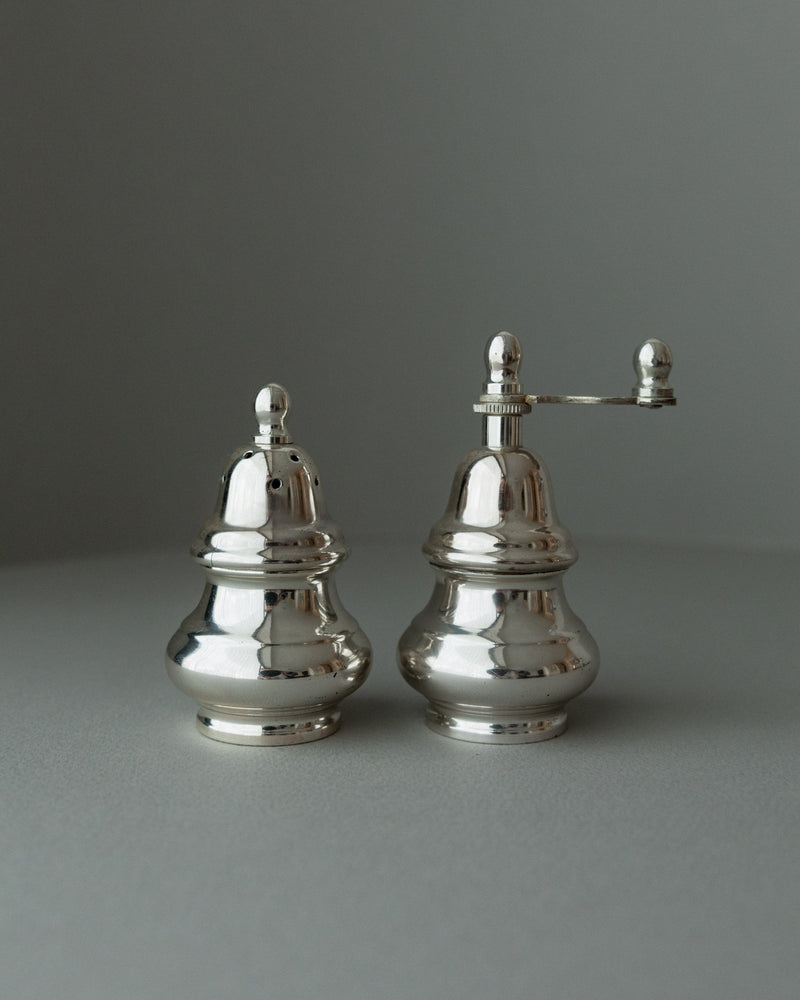 A pair of elegant, silver-plated Salt Shaker and Pepper Set - Italy 1970s by Bottega Jacobs sits on a neutral gray surface. The right shaker, vintage-inspired, features a small grinding handle, with lighting accentuating their shiny metallic finish.