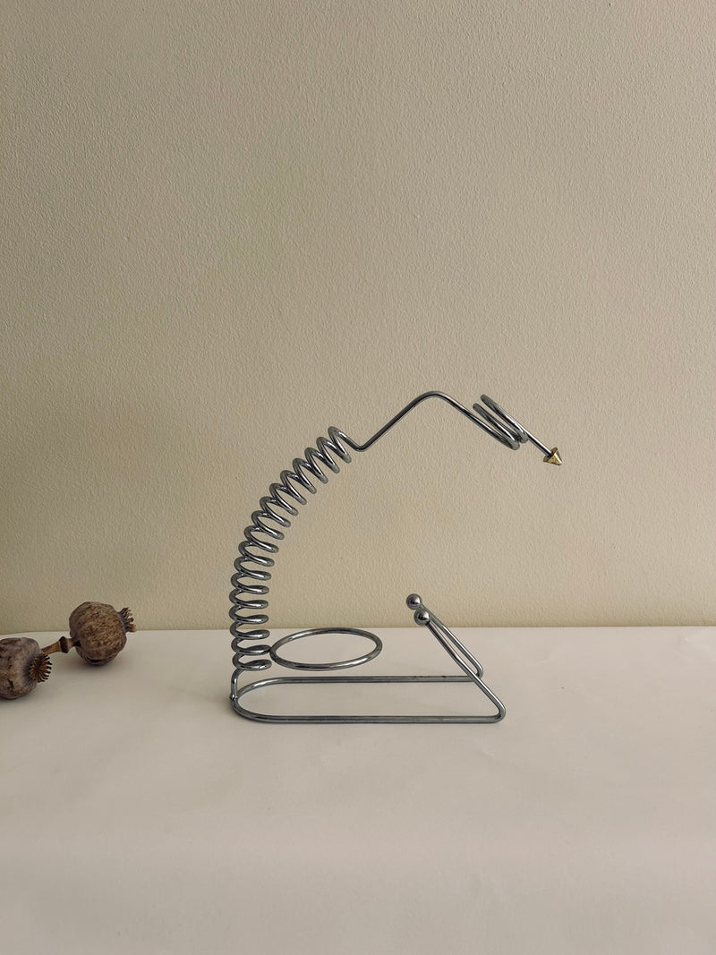 A sleek metal spiral resembling a coiled spring with a triangular tip, part of the minimalist design of Flock Studio's Postmodern Wine Holder, stands against a plain beige background. Two dried seed pods lie on the left side of the white surface, blending art with functionality.