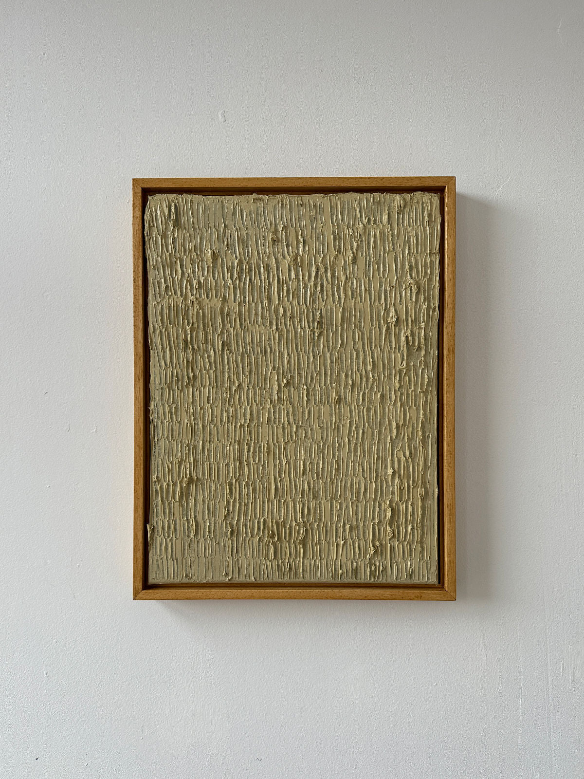 The "Buff Titanium" painting by Roi Elmaliah, featuring a textured surface and set in a wooden frame, is mounted on a white wall. Its beige textured ridges evoke the rustic charm of Amsterdam, giving it a tactile, layered appearance.