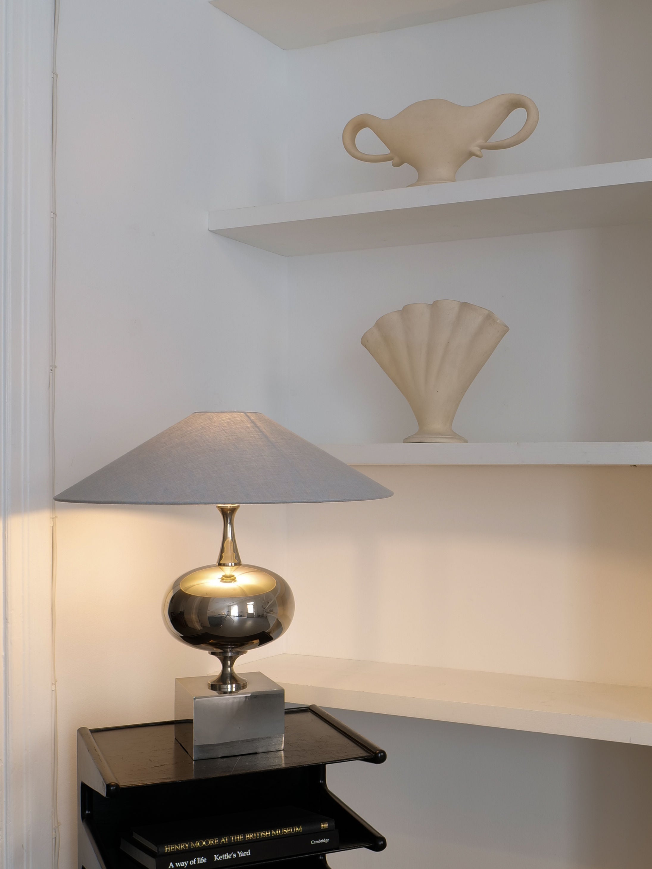 A decorative shelf with minimalist design elements features the Barbier Sculptural Lamp 1970s from Collection apart, adorned with a gray lampshade typical of 1970s French lighting. Above it, two art pieces enhance the scene: a cream-colored sculptural vase shaped like a trophy and a fan-shaped ceramic piece. The overall ambiance is calm and elegant.