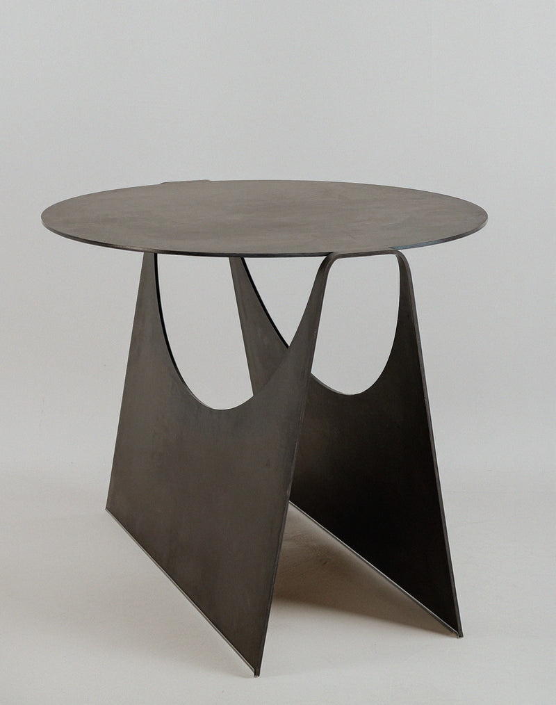 The 'Maru' Side Table by Modan features a modern, minimalist design with a round steel sheet top and two triangular supports with large semicircular cutouts, creating a distinctive look against a plain white background.