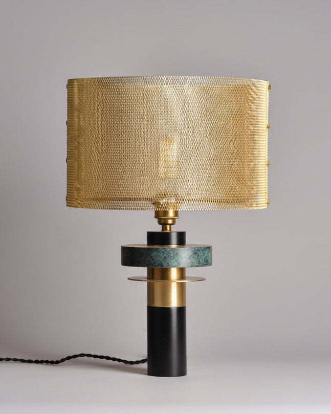 A modern Dyane table lamp by Marine Breynaert featuring a cylindrical base with mixed textures in black and verdigris, topped with a golden deployed stainless steel lampshade that encases a visible bulb.